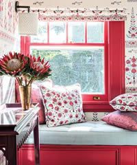 Window seat with red frame, floral pillows, and patterned wallpaper.