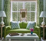 Green couch with patterned pillows and lamps, in front of a window with a chair.