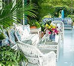 White wicker furniture with floral cushions on a sunlit porch, surrounded by lush greenery and flowers.