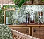 Rattan bar setup with green and white patterned wallpaper, featuring a cocktail shaker, bottles, and a lamp.