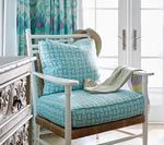 White armchair with aqua cushions, patterned pillow, and folded blanket; window in background.