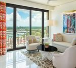 Living room with floor-to-ceiling windows, cityscape view, white sofa, patterned rug, armchairs, and colorful abstract art.