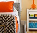 Bed with orange pillow and patterned blanket next to a white nightstand with a lamp.