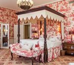 Bedroom with red toile wallpaper, canopy bed, patterned bench, and chandelier.