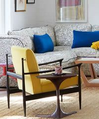 Yellow chair beside a patterned couch with blue pillows, next to a small table.