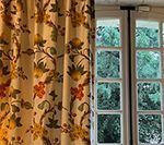 Cream-colored curtain with floral pattern beside a white-framed window. Green foliage visible through window panes.