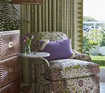 An armchair with patterned fabric, purple pillow, side table, and chest of drawers, next to striped curtains and an outdoor view.