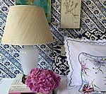 White lamp with beige shade on a table, next to pink flowers and a bed with a floral pillow.