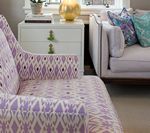 A patterned lavender armchair next to a white nightstand and a light gray sofa with decorative pillows.