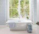 White bathtub in front of a large window, blue patterned trash can on the right.
