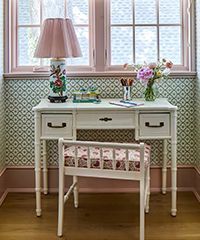 Desk with lamp and floral wallpaper near a window. Stool in front.