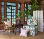 Sunroom with wicker chair, floral armchair, small table, pink pillows, and tall windows.