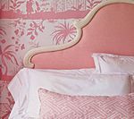 Pink upholstered bed headboard against matching patterned wallpaper, white bedding.