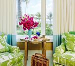 Two floral green armchairs frame a window with a small table and a vase of pink flowers.