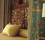 A bed with a carved wooden headboard and yellow pillows, beside a teal floral patterned wall.