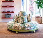Round patterned sofa with three pillows, green base, in a shop setting with shelves and a window.