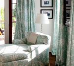 Chaise lounge by a window with light blue patterned curtains, white lamp, framed photos.