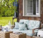 Outdoor seating area with blue and white floral cushions, wicker tables, and greenery backdrop.
