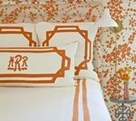 Bed with white and orange bedding, monogrammed pillows, and patterned wallpaper.