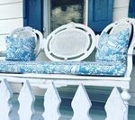 White bench with blue cushions and pillows on a porch, white picket fence in front.