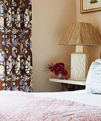 Bedroom corner with floral curtains, lamp, flowers, and a bed with a pink blanket.