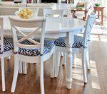 White dining set with round table, cross-back chairs with patterned cushions, in sunlit room.