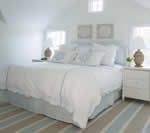 White bedroom with king-size bed. Light blue and tan striped rug, white furniture, two lamps and artwork.