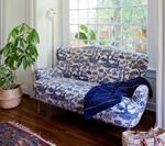 Blue and white patterned loveseat by a window, with a blanket draped on it. A houseplant sits nearby.