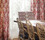 Red patterned curtains frame a sunny window with a woven-backed chair and dining table.