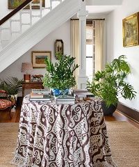 Round table with patterned cloth, potted plants, books, and staircase in a bright hallway.