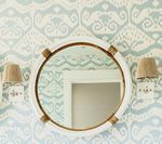 Round white mirror with brass accents, flanked by sconces, against patterned blue and white wallpaper.