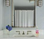 Bathroom with a white countertop, silver faucet, and mirror reflecting a white shower curtain. Blue flowers and a pink bottle sit on the counter.