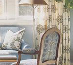 A light blue sitting area with an upholstered chair, sofa, patterned curtains, and a floor lamp.