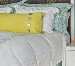 Bed with white and teal patterned pillows, a yellow bolster pillow, and white bedding.