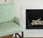 Green patterned armchair beside a white marble fireplace with logs.