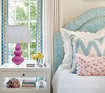 Bedroom with light blue patterned headboard, white bedding, and pink lamp on a nightstand with blue patterned curtains.