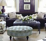 Living room with purple accent wall, dark sofa, patterned ottoman, and floral accents.