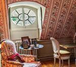 Room with patterned wallpaper, oval window, ornate furniture, and a small table with items.
