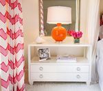 White nightstand with orange lamp and pink flowers. Pink chevron curtains on the left.