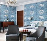Bedroom with blue patterned wallpaper, two gray armchairs, a wooden dresser, and a bed.