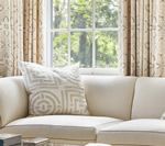 Cream-colored sofa with patterned pillow in front of a window with beige curtains.