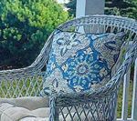 Blue patterned pillow on a gray wicker chair on a porch, green foliage in the background.