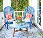 Two blue wicker chairs on a porch with cushions, round table, and plants.