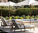Lounge chairs under umbrellas beside a pool and manicured hedge.