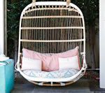 A hanging wicker chair with pillows. Pink and white cushions. Outdoors, near a teal object.