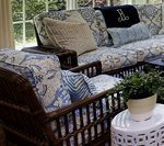 Wicker furniture with blue and white patterned cushions, cream pillows, and a black monogrammed throw, next to a white side table.