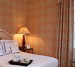 Bedroom with patterned wallpaper, lamp, bed, tray of cups, and patterned curtain.