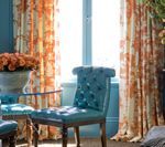 Teal upholstered chair in a room with orange floral curtains and a round glass table.