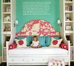 A child sits on a white bed with a floral headboard, teal wall, and built-in bookshelves.