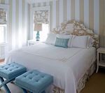 Bedroom with striped wallpaper, bed with a floral headboard, and blue stools at the foot of the bed.